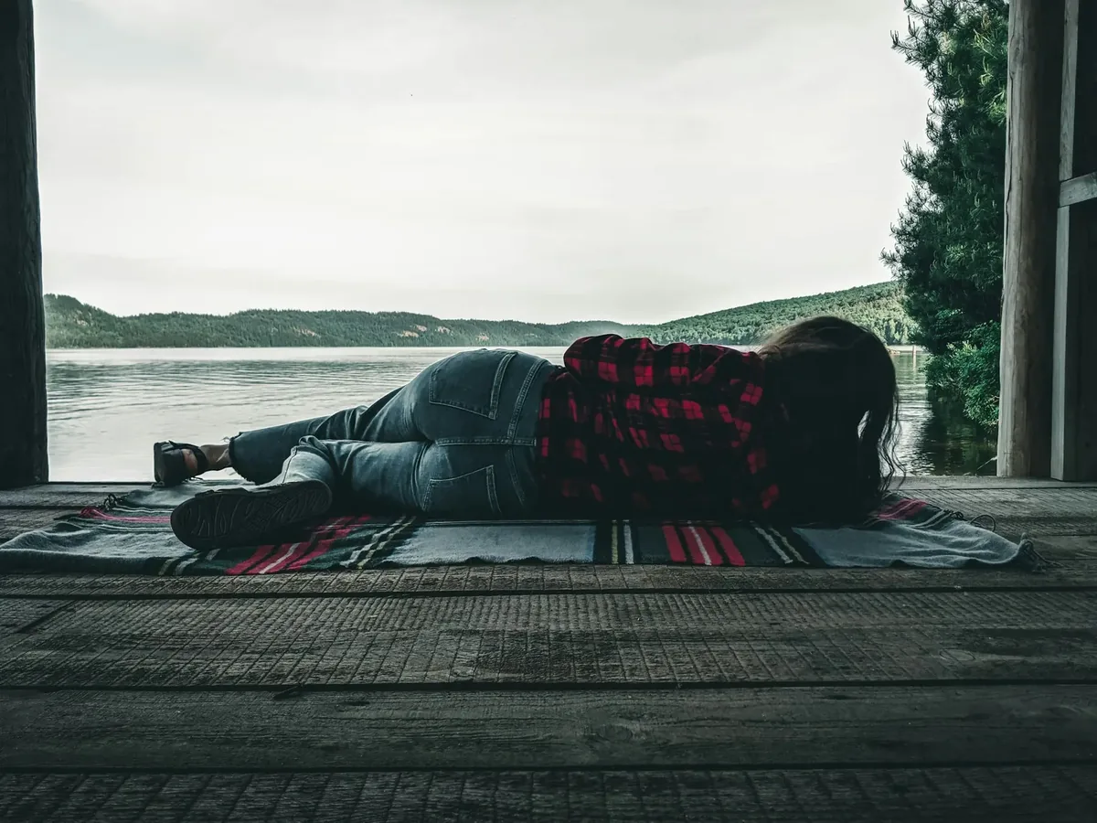 A person lying at rest on a rug, facing a quiet lake